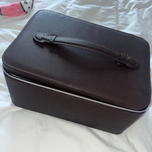 Brun necessär från CAIA i skinn - Snygg och stilren brun necessär från CAIA i skinn med silverfärgad dragkedja och praktiskt handtag upptill. Perfekt för att organisera smink eller småprylar när du är på språng. Rektangulär form och diskret logga på framsidan.