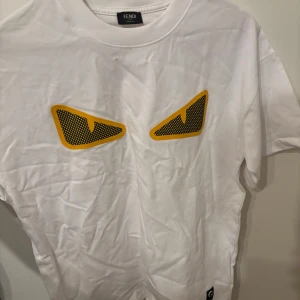 Vit Fendi t-shirt med ögonprint - Säljer en vit t-shirt från Fendi med ikoniska gula och svarta ögonprint på bröstet. T-shirten har korta ärmar och en rund hals. Detalj med svart tygpatch och knapp på ärmen, samt Fendi-logga i nacken. Tillverkad i mjuk bomull.