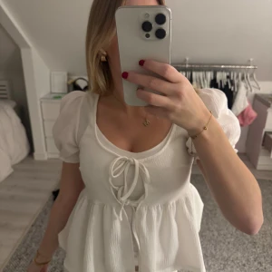 Vit blus med puffärm och knyt - Superfin vit blus med puffärmar och två knyt framtill. Blusen har en lätt rynkad struktur och volang nedtill som ger en söt och trendig look. Perfekt för dig som gillar romantiska detaljer och vill ha en luftig topp med feminin touch.