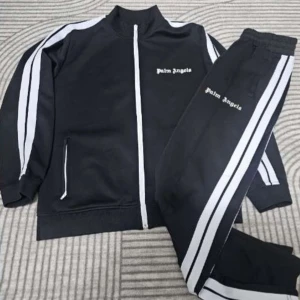 Palm Angels svart  - Svart träningsset från Palm Angels med vita ränder längs ärmar och ben. Setet består av en zip-jacka med ståkrage och matchande byxor, båda med Palm Angels-logga i vitt. Materialet är mjukt och stretchigt, perfekt för chill eller sport.
