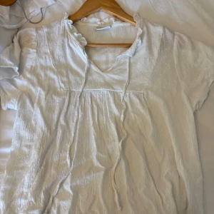 Vit blus med snörning och volang - Säljer en söt vit blus med korta ärmar, v-ringning och snörning framtill. Blusen har volangdetaljer vid halsen och är gjord i ett lätt, krinklat bomullstyg som ger en chill och luftig känsla. Perfekt för ljusa dagar och enkel att styla.