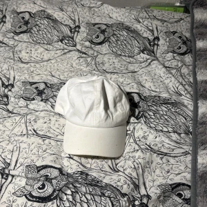 Vit klassisk keps med justerbart spänne - Säljer en stilren vit keps med böjd skärm och justerbart metallspänne bak. Kepsen är tillverkad i bomull och har en enkel, clean look som passar till många outfits. Perfekt för dig som gillar minimalistisk stil och vill ha en keps som funkar året runt.