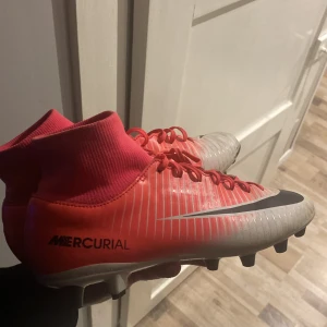 Nike Mercurial fotbollsskor rosa - Säljer ett par Nike Mercurial fotbollsskor i en snygg rosa och vit färg med svarta detaljer. Skorna har snörning, hög strumpliknande krage och dobbar för gräsplan. Perfekta för dig som vill sticka ut på planen och få grymt grepp. Materialet är syntet.
