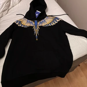 Marcelo Burlon svart hoodie med vingar - Svart hoodie från Marcelo Burlon County of Milan med ikoniskt vingtryck i blått och gult över axlar och bröst. Tröjan har huva med snörning och stor känguruficka framtill. Perfekt för dig som vill sticka ut med en fet streetwear-look.