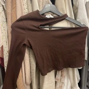 Brun ribbad croppad topp från Bershka - Säljer en brun croppad topp från Bershka med ribbad struktur och cool cut-out detalj vid halsen. Toppen har lång ärm och är figurnära, perfekt för dig som gillar trendiga och unika plagg. Materialet är mjukt och stretchigt för skön passform.
