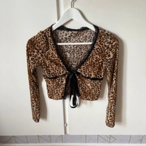 Leopardmönstrad croppad bolerotopp Shein - Säljer en croppad bolerotopp från Shein i leopardmönster med svarta spetsdetaljer och satinband som knyts framtill. Toppen har lång ärm och v-ringad hals. Materialet är mjukt och stretchigt, perfekt för dig som gillar att sticka ut med djurmönster.