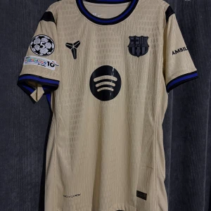 FC Barcelona Spelarnas version 25/26 – Raphinha #11 – Champions League Edition - Barcelona bortatröja från säsongen 2025/26 i guld färg, med Raphinha #11 på ryggen.  Champions League-version med officiella patchar och sponsorloggor (Spotify, UNHCR, Ambilight TV). Nike Dri-FIT ADV-material – mycket bekväm och ventilerande.  Skick: Helt ny med prislapp   Perfekt för samlare eller Barça-fans! 💙❤️