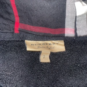 Svart rutig hoodiejacka Burberry - Svart hoodiejacka från Burberry med klassiskt rutigt foder i svart, vitt och rött. Jackan har snörning i huvan med metalländar och dragkedja framtill. Materialet är bomull och elastan, vilket gör den mjuk och bekväm. Perfekt för dig som gillar stilren design med lyxig känsla.