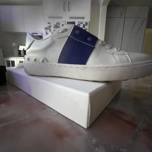 Valentino Garavani vita sneakers med blå detalj - Snygga vita sneakers från Valentino Garavani med bred blå rem över sidan och coola nitar på hälen. Skorna har snörning och platt sula, tillverkade i skinn för en lyxig känsla. Perfekt för dig som gillar stilrena och exklusiva sneakers. Storlek42 men passar även 43