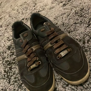 Svarta sneakers från Hugo Boss - Snygga svarta sneakers från Hugo Boss med bruna detaljer och snörning. Skorna har en kombination av mocka och textil, samt en beige sula. Märkeslogga syns både på sidan och framtill vid snörningen. Perfekta för dig som gillar stilrena och klassiska sneakers.