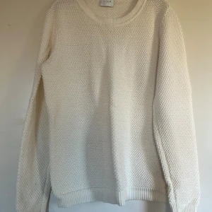 Vit stickad tröja från Vila - Klassisk vit stickad tröja från Vila med rund halsringning och lång ärm. Tröjan har en grovstickad struktur och ribbade muddar vid ärmslut och nederkant. Perfekt för lager-på-lager under kyliga dagar.