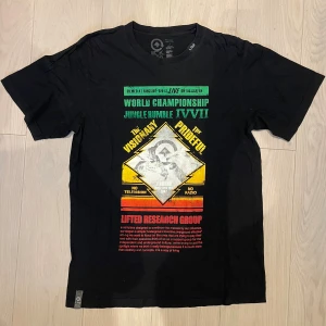 Svart t-shirt från Lifted Research Group - Cool svart t-shirt från Lifted Research Group med färgstarkt tryck i grönt, gult och rött framtill. Motivet har texten 'World Championship Jungle Rumble' och grafiska detaljer. 