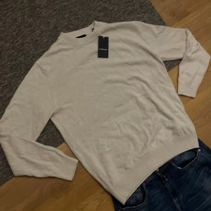 Beige stickad tröja från Lindbergh - Snygg beige stickad tröja från Lindbergh i storlek S, men stor i storleken så passar M. Tröjan har rund hals, ribbade muddar och ett diskret broderat emblem framtill. Perfekt för dig som gillar stilrena och klassiska plagg med en modern touch, Perfekt till den kalla vintern. Ny pris 999kr (syns i bild nmr 2)