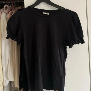 Svart topp med puffärm - Säljer en svart topp med puffiga korta ärmar och söt volangdetalj vid ärmslutet. Toppen har rund halsringning och är gjord i mjukt bomullsmaterial. Perfekt för dig som gillar stilrena och enkla plagg med en twist.