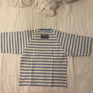 Randig t-shirt från Mads Nørgaard - Säljer en fräsch t-shirt från Mads Nørgaard med breda, ljusblå och vita ränder. T-shirten har korta ärmar och en rund, ribbad halsringning i ljusblått. Märkets logga är broderad i brunt på ryggen nära nacken. Perfekt för dig som gillar clean och skandinavisk stil. Sååå unik också!!💙🤍 alldrig använt tröjan.