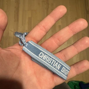 Blått Christian Dior J'Adior armband - Snyggt blått armband från Christian Dior med texten 'J'Adior' broderad i vitt. Armbandet har en vävd design med ljusblå och vita detaljer samt knytning med tofsar. Perfekt accessoar för dig som vill ha en lyxig och trendig touch på din outfit.