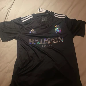 Svart Real Madrid x Balmain tröja Adidas - Säljer en svart fotbollströja från Adidas i samarbete med Real Madrid och Balmain. Tröjan har korta ärmar, silvriga detaljer och klassiska Adidas-ränder på axlarna. Materialet är lätt och andas, perfekt för träning eller match. Pris kan diskuteras 