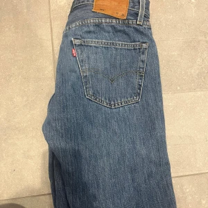 Levi's 501 blå jeans straight fit - Klassiska blå Levi's 501 jeans med rak passform och femficksdesign. Jeansen har knappgylf, tydliga kontrastsömmar och den ikoniska läderpatchen bak i midjan. Tillverkade i slitstarkt bomullsjeansmaterial, perfekt för en avslappnad och tidlös look.