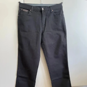 Svarta jeans från Wrangler 34/36 - Svarta jeans från Wrangler i 100% bomull med klassisk femficksdesign och raka ben. Byxorna har bälteshällor, knapp och dragkedja framtill samt Wrangler-logga på bakfickan. Perfekta för en clean och stilren look.