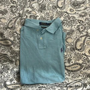 Ljusblå pikétröja från Polo Ralph Lauren - Snygg ljusblå pikétröja från Polo Ralph Lauren i custom slim fit. Klassisk krage, korta ärmar och två knappar framtill. Ikoniska lilla loggan i lila broderad på bröstet. Perfekt för dig som gillar stilren och enkel design. Det står L men sitter mer som en störrr M, har lite fläckar men lätt att få bort. Billigare vid snabb affär🤩