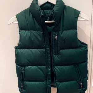 Grön dunväst barn från Polo Ralph Lauren - Mörkgrön dunväst från Polo Ralph Lauren (The Gorham Down Gilet) med hög krage och tre dragkedjefickor framtill i storlek S (8-10 år). Västen har klassisk quiltad design och diskret logga på bröstet. Perfekt att slänga över en hoodie eller tröja för extra värme. Inköpt på NK i Stockholm och väldigt sparsamt använd.  