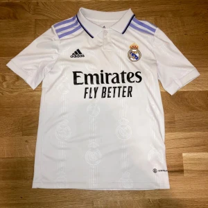 Real Madrid Jersey 22/23  - Säljer en officiell Real Madrid fotbollströja från Adidas i vit polyester med svarta tryck och broderat klubbmärke. Tröjan har korta ärmar, krage med knappar och lila ränder på axlarna. Mönster med Real Madrid-loggor i tyget och 'Emirates Fly Better' tryckt på bröstet.
