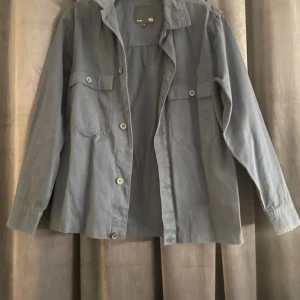 Grå overshirt från 157, storlek S - Stilren grå overshirt från 157 med klassisk krage, två bröstfickor med knappar och raka långa ärmar. Skjortan är i bomull och har en avslappnad passform, perfekt att slänga över en t-shirt. Enkel och clean look. Endast testad några gånger! 