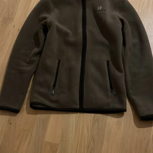 Brun fleecejacka från Chevalier - Mjuk och varm brun fleecejacka från Chevalier med hel dragkedja framtill och två sidofickor med dragkedja. Jackan har lång ärm och en enkel, clean design med diskret logga på bröstet. Perfekt för kyliga dagar och sköna promenader.