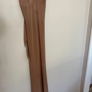 CLUB L London klänning - Säljer en snygg beige långklänning med smala axelband och v-ringning. Klänningen har ett coolt knyt i ryggen och är tillverkad i ett mjukt, slätt material som faller fint längs kroppen. Perfekt för dig som gillar minimalistisk stil och vill sticka ut med en enkel men elegant look. 