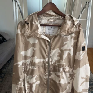 Beige camo vindjacka från Woolrich - Snygg beige och vit camouflage vindjacka från Woolrich med huva och dragkedja framtill. Passar nu till hösten för dig som vill sticka ut lite. Storlek M och är som nyskick. Använt varsamt. För fler frågor eller funderingar är det bara att höra av sig 