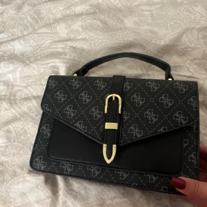 Svart handväska  - Kan sänka pris Snygg svart handväska  med klassiskt monogrammönster och guldfärgade detaljer. Väskan har ett stilrent spänne framtill, praktiskt handtag och flera fack inuti, inklusive dragkedjefack. Tillverkad i syntetmaterial med elegant finish.