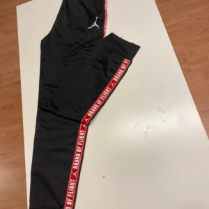 Jordan Brand of Flight svarta byxor XL - Svarta byxor från Jordan med röd resår i midjan och röda sidor med texten 'Brand of Flight'. Byxorna har vit Jumpman-logga på låret och är tillverkade i polyester. Perfekta för en sportig och avslappnad stil. För längd 163-175