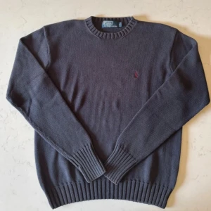 Mörkblå stickad tröja Polo Ralph Lauren Vintage - Mörkblå stickad tröja från Polo Ralph Lauren i 100% bomull. Klassisk modell med rund halsringning, ribbade muddar och diskret röd logga broderad på bröstet. Perfekt för dig som gillar stilrena och tidlösa plagg. 