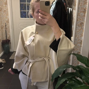 Beige kappa med bälte och hög krage - Säljer en beige kappa med hög krage och snyggt bälte i midjan. Jackan har vida ärmar och stängs med en guldig knapp upptill. Perfekt för dig som vill ha en stilren och trendig look. Materialet känns mjukt och har en lyxig känsla.