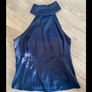 Mörkblå halterneck topp  - Snygg mörkblå halterneck topp i glansigt satintyg. Toppen har hög krage med knappar i nacken och dragkedja i sidan. Den är ärmlös och har en elegant, figurnära passform som ger en stilren look. Knapparna är satta av mig men märks inte 