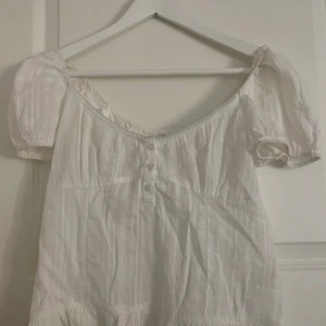 Vit blus med volang och knappar - Söt vit blus med kort puffärm, knappar framtill och volangkant nertill. Blusen har en lätt rynkad struktur och subtila ränder i tyget. Perfekt för dig som gillar romantisk och luftig stil.