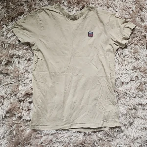 Beige t-shirt från Vinson, storlek L - Beige t-shirt från Vinson i storlek L. Klassisk modell med rund hals och korta ärmar. Liten broderad logga på bröstet med röd, vit och blå detalj. Tillverkad i mjuk bomull som är skön mot huden.