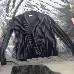 Svart ribbad kofta från Freequent - Svart kofta från Freequent i storlek S. Den har ett omlottliknande fall framtill och är ribbad i materialet, vilket ger en snygg struktur. Långärmad och i mjuk stickad kvalitet, perfekt för lager på lager. Enkel att matcha med olika outfits.