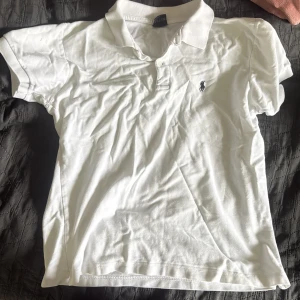 Vit pikétröja från Ralph Lauren - Vit pikétröja från Ralph Lauren med klassisk krage, korta ärmar och knappar framtill. Ikoniska broderade loggan på bröstet. Tillverkad i mjuk bomull för en skön och avslappnad stil.