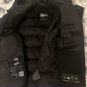 Mörkblå dunjacka Sail Racing med huva - Säljer en mörkblå dunjacka från Sail Racing med huva och dragkedja. Jackan är vadderad med 600 fill power dun och har två sidofickor med dragkedja. Perfekt för kalla dagar och har en stilren look med logga på bröstet och huvan.