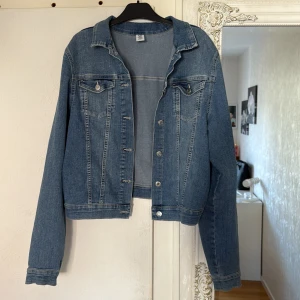Blå jeansjacka från H&M, strl 170 - Klassisk blå jeansjacka från &H&M med raka linjer, bröstfickor med knappar och silverfärgade detaljer. Jackan har en normal passform och är tillverkad i bomull med lite polyester och elastan för extra komfort. Perfekt till din vardagsstil.