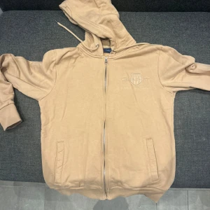 Beige hoodie från Gant med dragkedja - Säljer en beige hoodie från Gant med broderad logga på bröstet. Tröjan har huva med snörning, hel dragkedja framtill och två fickor. Tillverkad i mjuk bomull och har långa ärmar med ribbade muddar. Perfekt för chill dagar.