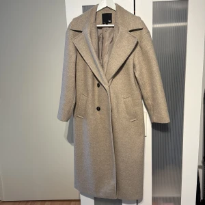 Beige kappa från 157 i ullmix XS - Stilren beige kappa från 157 i ullmix med klassisk krage, två knappar framtill och fickor på sidorna. Lång modell med snyggt fall och helfodrad insida. Perfekt för dig som gillar en clean och tidlös look.