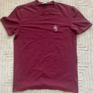 Vinröd t-shirt från Dolce & Gabbana - Vinröd t-shirt från Dolce & Gabbana med broderad DG-logga i vitt på bröstet. Klassisk rund hals och korta ärmar. Storlek M, vid fler frågor eller funderingar är det bara att skriva!