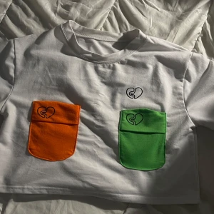 Vit croppad topp med färgglada fickor - Vit croppad topp i 100% bomull med två stora fickor framtill, en orange och en grön, båda med svart hjärtdetalj. Rund halsringning och kort ärm. Perfekt för dig som gillar färg och unika detaljer.