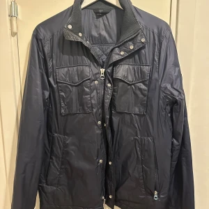 Mörkblå quiltad jacka från J.Lindeberg - Snygg mörkblå quiltad jacka från J.Lindeberg med dragkedja och tryckknappar. Jackan har två bröstfickor med lock, sidfickor med dragkedja och en hög krage. Tillverkad i tekniskt material från Olmetex och fodrad med Thermore.