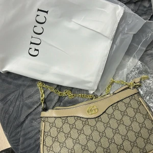 Gucci axelväska med GG-mönster - Snygg axelväska från Gucci med klassiskt GG-mönster i beige och brunt. Väskan har guldfärgade detaljer, kedjerem och bruna skinnpartier. Perfekt för dig som vill ha en ikonisk och trendig accessoar med lyxig känsla.