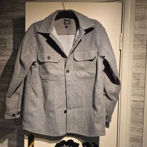 Grå overshirt från H&M med fickor - Stilren grå overshirt från H&M med klassisk krage, två stora bröstfickor med lock och knappar samt knappar vid ärmslut. Jackan är i ullblandning och har en rak, något loose passform. Perfekt lager-på-lager-plagg för höst och vår.