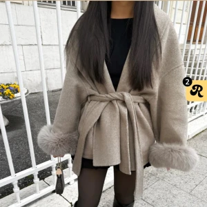 Beige kappa med päls och bälte - Helt ny beige kappa fortfarande stängd i förpackningen med breda slag, knytskärp i midjan och fluffiga pälsdetaljer vid ärmsluten. Jackan har en loose passform och är perfekt för dig som vill ha en trendig och mysig look under höst och vinter.