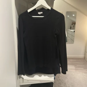 Svart långärmad tröja från Gina Tricot - En stilren svart långärmad tröja från Gina Tricot. Tröjan har rund halsringning och är tillverkad i ett mjukt, ribbat material som sitter skönt mot kroppen. Perfekt att matcha med jeans eller kjol för en enkel och snygg look.